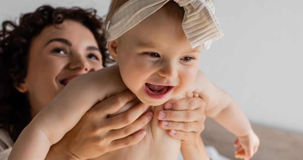 curly woman holding happy infant daughter in headband with bow, banner