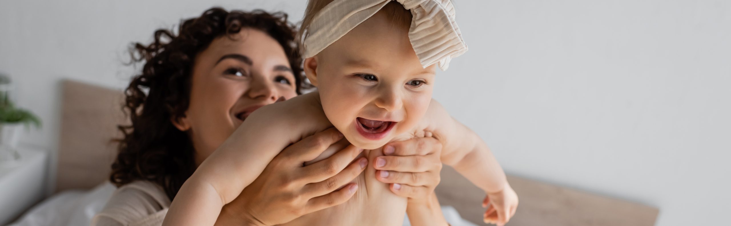 curly woman holding happy infant daughter in headband with bow, banner